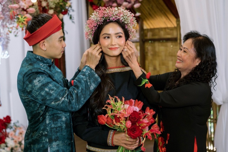 Tuấn Khôi và mẹ chồng trao trang sức cho H'Hen Niê. Cô dâu đi chân đất khi diện trang phục truyền thống của người Ê-Đê và đội vòng hoa baby với tông trắng, hồng chủ đạo.