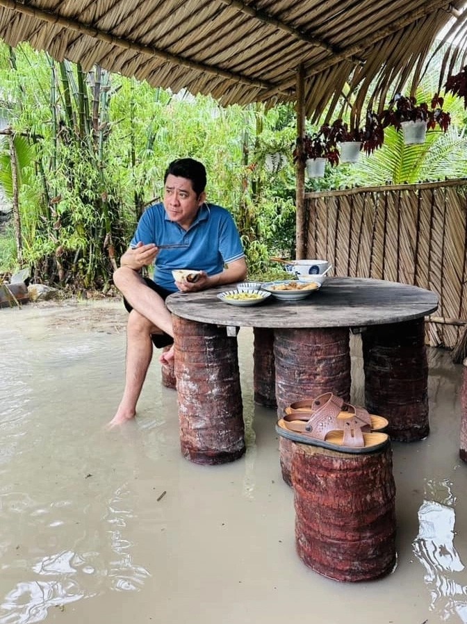 Huỳnh Anh Tuấn ăn cơm trong nhà chòi dựng giữa khu đất 6.000 m2 trang bị đơn sơ, hồi tháng 6. Sau mưa lớn, nền nhà bị nước ngập tới mắt cá chân.